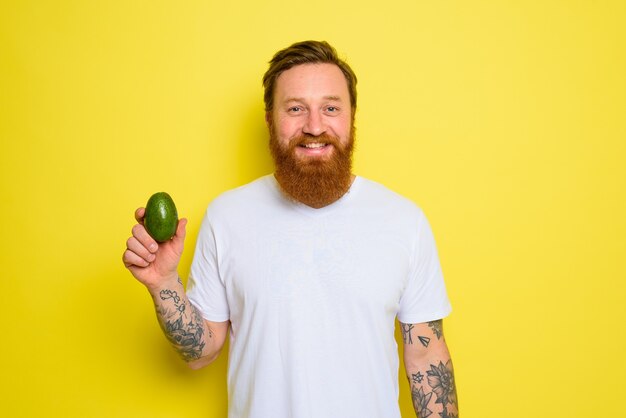 Hombre feliz con barba y tatuajes sostiene un aguacate