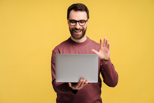 Hombre feliz, barbudo, independiente, hipster, con gafas, sosteniendo una computadora portátil, saludando en una llamada de video