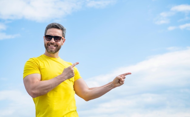 Un hombre feliz con camisa amarilla y gafas de sol señala con el dedo sobre el fondo del cielo con espacio para copiar