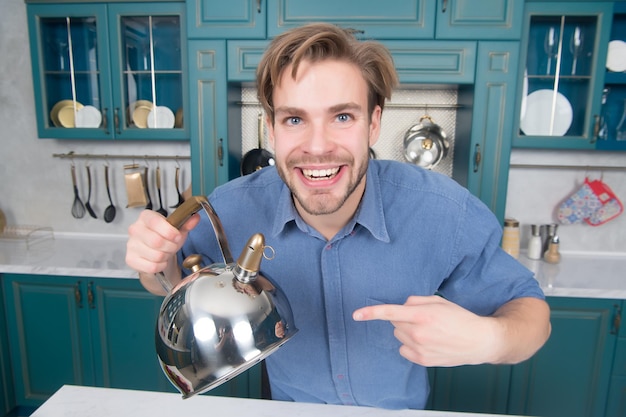 Hombre feliz en camisa azul señalar con el dedo a la tetera. Electrodomésticos y muebles de cocina. Preparación de alimentos, cocción, calentamiento, agua hirviendo. Hora del té, humor del té. Presentación de producto, venta online.