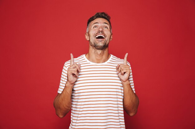 Hombre feliz en camiseta a rayas gesticulando los dedos índices hacia arriba en copyspace aislado en rojo