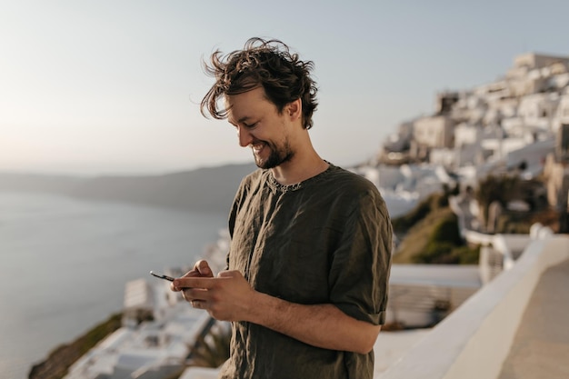 Un hombre feliz con una camiseta verde oscuro sonríe ampliamente y sostiene el teléfono. Un chico moreno genial posa de buen humor en un lugar con vistas al mar y a la ciudad.