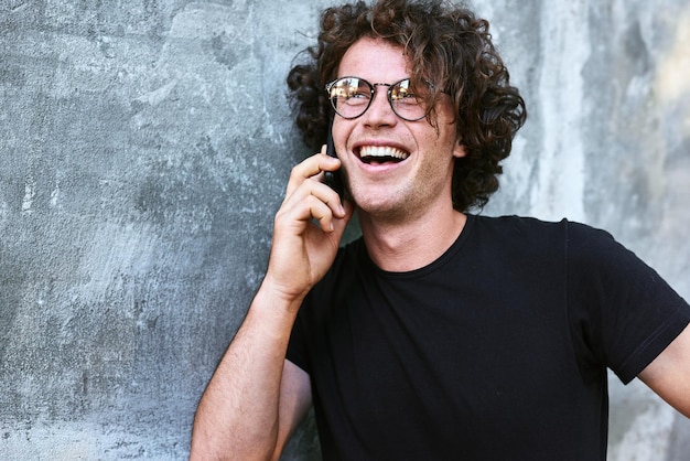 Hombre feliz caucásico guapo de pie al aire libre hablando por teléfono móvil Hombre joven con cabello rizado usa gafas descansando afuera en la ciudad haciendo una llamada en su teléfono celular sobre fondo de hormigón