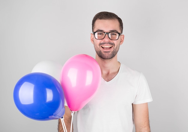 Hombre feliz celebra el aniversario de una empresa comercial, viene a una fiesta corporativa, lleva globos de colores, usa gafas, una camiseta blanca, posando en la oficina