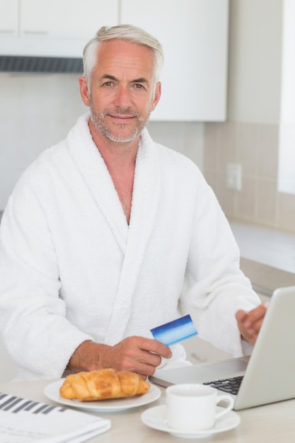 Hombre feliz de compras en línea en el desayuno en una bata de baño