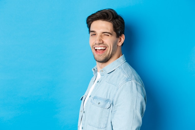 Hombre feliz y confiado gire la cabeza a la cámara, guiñando un ojo y sonriendo, de pie sobre fondo azul.