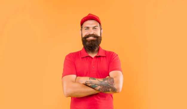 Hombre feliz y confiado con gorra roja casual y camiseta manteniendo brazos tatuados cruzados con fondo amarillo repartidor