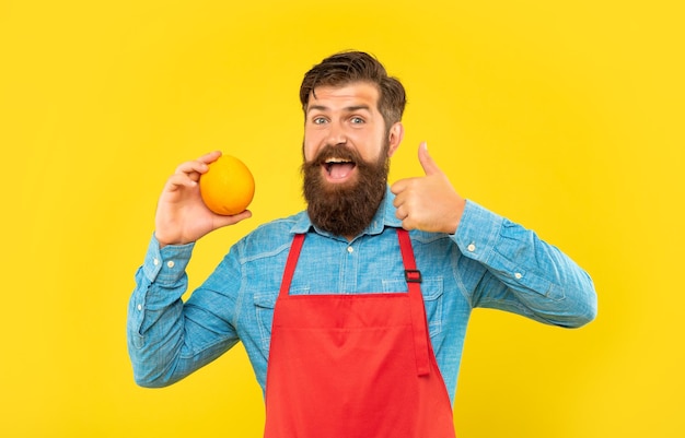 Hombre feliz en delantal dando pulgar a fruta cítrica naranja fresca fondo amarillo frutero
