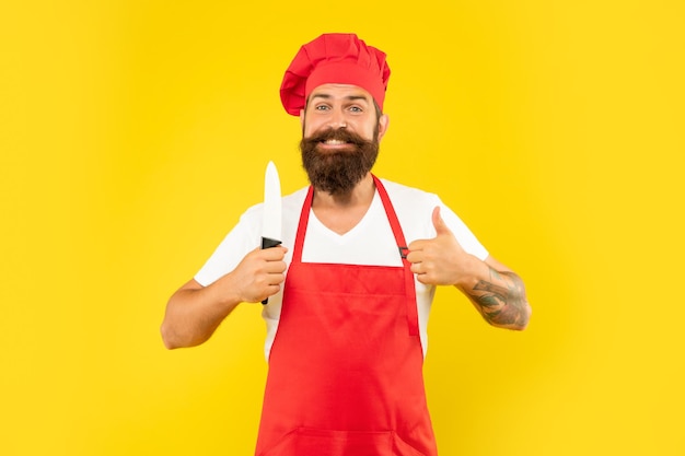 Hombre feliz en delantal rojo y toque dando pulgar sosteniendo chefs cuchillo fondo amarillo, chef