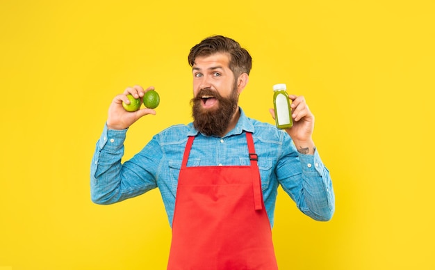 Hombre feliz en delantal sosteniendo limas y botella de jugo de fondo amarillo, barman de jugo.