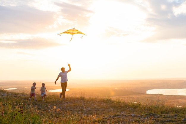 Hombre feliz y dos niños, padre e hijos, corren con cometas en la naturaleza al atardecer