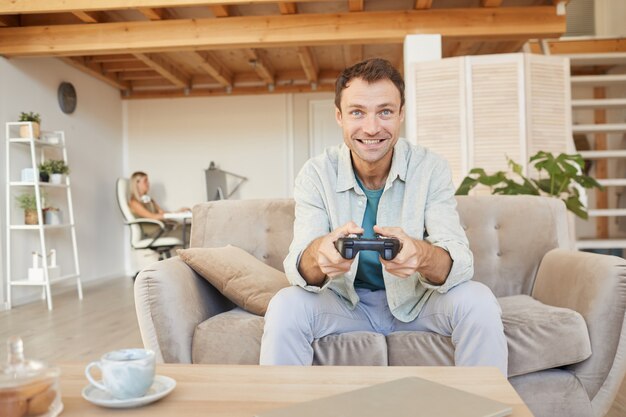 Hombre feliz emocionado sentado en el sofá con joystick y jugando juegos de computadora en la sala de estar