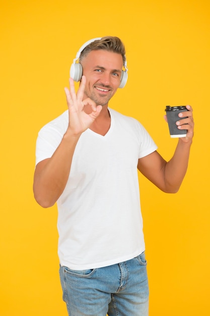 El hombre feliz escucha música que muestra el gesto de la mano OK para tomar café en un vaso desechable fondo amarillo coffeetogo