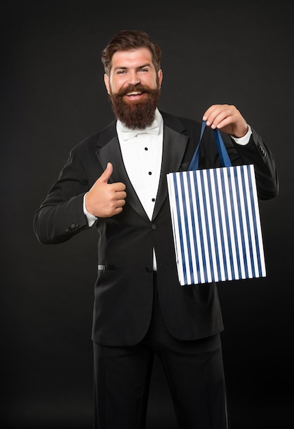 Hombre feliz en esmoquin pajarita ropa formal sobre fondo negro con compra bolsa copia espacio pulgar hacia arriba