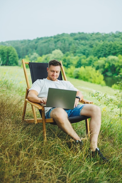 Un hombre feliz está relajándose en la naturaleza y trabajando remotamente en una computadora portátil Trabajo remoto mientras está de vacaciones Trabajando en una computadora portátil mientras está sentado en una silla en la naturaleza