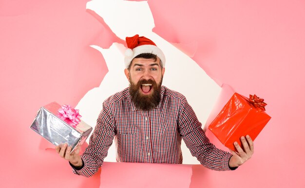 Hombre feliz con gorro de Papá Noel con regalos caja de regalos rompe a través de la pared de papel Navidad año nuevo barbudo