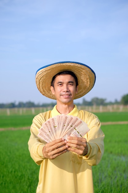 El hombre feliz del granjero usa la sonrisa de la camisa amarilla y sostiene el billete tailandés en la granja de arroz verde.