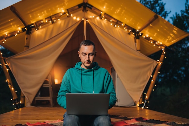 Hombre feliz independiente usando una computadora portátil en una acogedora carpa glamping en la noche de verano Tienda de campaña de lujo para vacaciones de verano al aire libre y concepto de estilo de vida de vacaciones