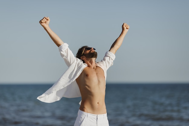 Hombre feliz levantando los brazos con camisa blanca en la playa