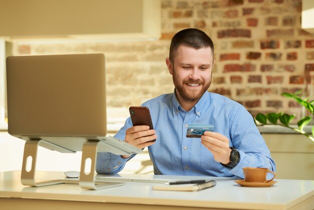 Un hombre feliz leyendo información del reverso de la tarjeta de crédito y escribiéndola en el teléfono inteligente para hacer una compra en línea en casa