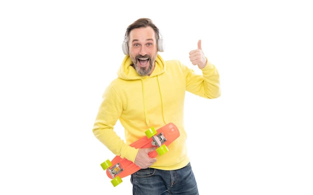 Hombre feliz maduro con auriculares y tablero de centavo chico mayor escuchar música aislado en blanco hombre en sudadera con capucha amarilla con patineta skater adulto estilo casual hipster skateboarding pulgar arriba