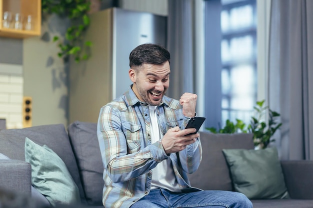 El hombre feliz mira el teléfono en casa y se regocija del éxito y la victoria.