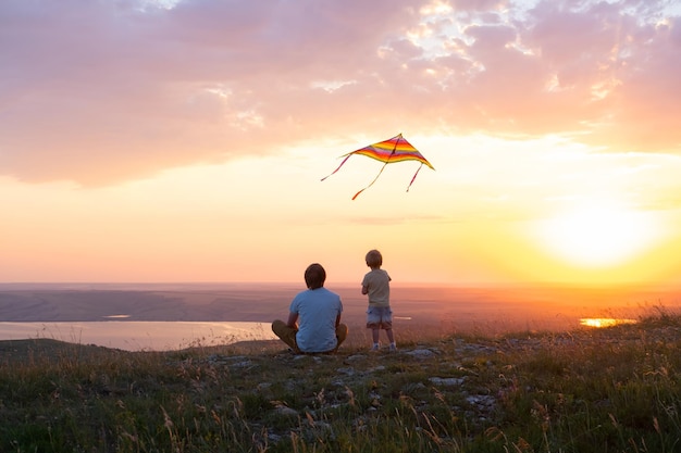 Hombre feliz y niño padre e hijo divirtiéndose con cometa en la naturaleza al atardecer