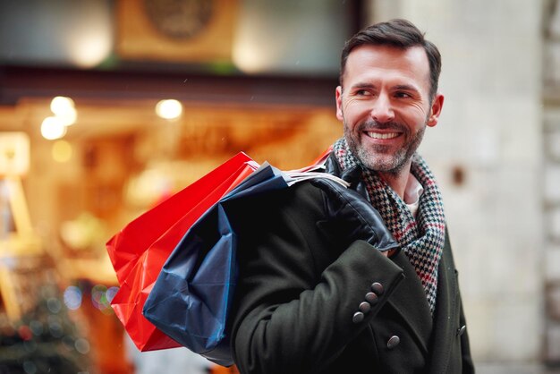 Hombre feliz y pensativo sosteniendo bolsas de compras mientras está de pie en la ciudad durante el invierno