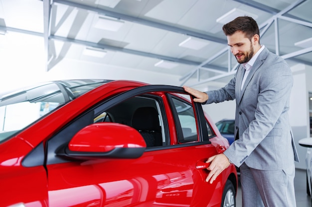 Hombre feliz de pie junto a un coche nuevo en el salón del automóvil y la puerta de apertura.