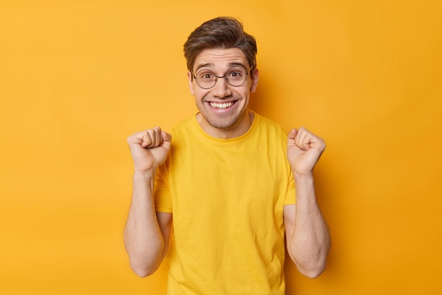 El hombre feliz y positivo aplaude por algo que aprieta los puños y se regocija de la victoria, sonríe con dientes y se siente muy contento de usar gafas redondas y una camiseta casual aislada sobre un fondo amarillo Triunfo y suerte