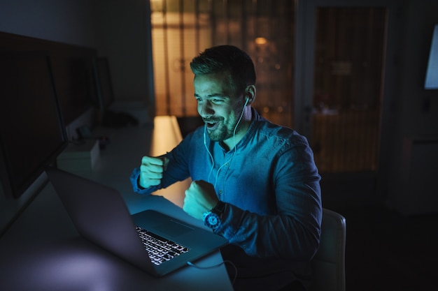 Hombre feliz que tiene videollamada y gesticulando wining mientras está sentado en la oficina a altas horas de la noche.