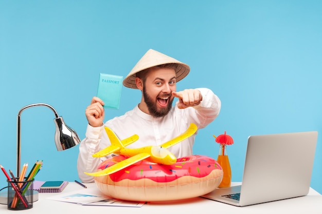 Un hombre feliz y relajado con barba en un sombrero divertido apuntándote con el dedo sosteniendo un pasaporte recomendando una gira caliente en el extranjero salvavidas y un avión de juguete tirado en el lugar de trabajo Foto de estudio interior aislada en fondo azul