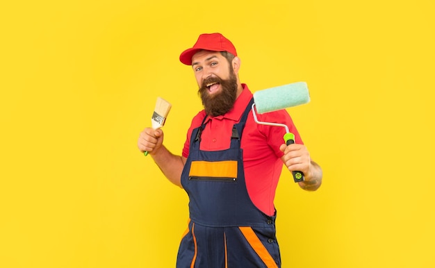 Hombre feliz en ropa de trabajo sostenga rodillo de pintura y cepillo sobre fondo amarillo