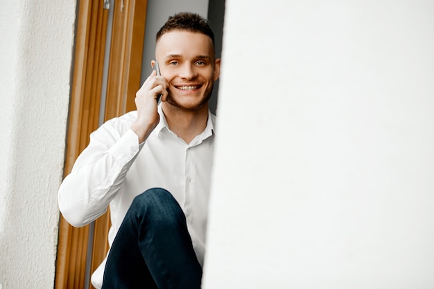 Hombre feliz sentado en el alféizar de la ventana y hablando por teléfono