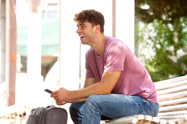 Hombre feliz sentado en el banco del parque con la maleta y el teléfono celular