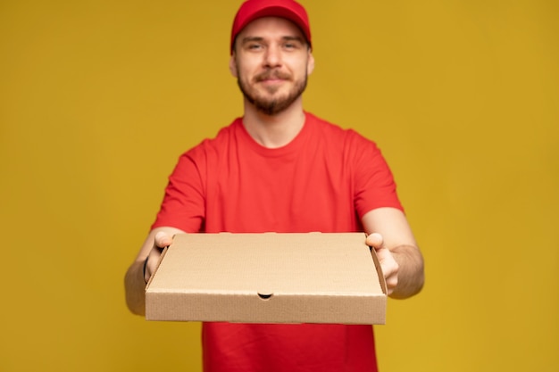 Hombre feliz del servicio de entrega en camiseta roja y gorra dando orden de comida y sosteniendo una caja de pizza aislada sobre una pared amarilla