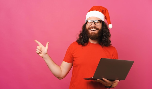 El hombre feliz con sombrero de Navidad señala a un lado y sostiene una computadora portátil
