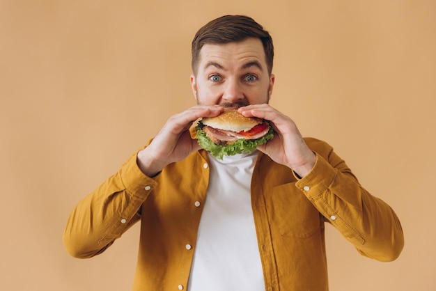 Hombre feliz y sonriente con barba en camisa amarilla comiendo hamburguesa sobre un fondo beige