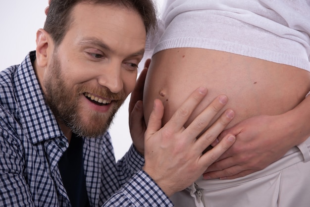 Foto el hombre feliz y sonriente está escuchando el vientre embarazado de la mujer.