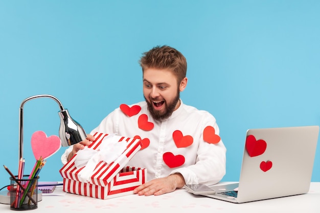 Hombre feliz sorprendido con barba en camisa blanca en corazones rojos pegajosos desempaquetando el presente y mirando dentro con una sonrisa satisfecha con el regalo Disparo de estudio interior aislado sobre fondo azul