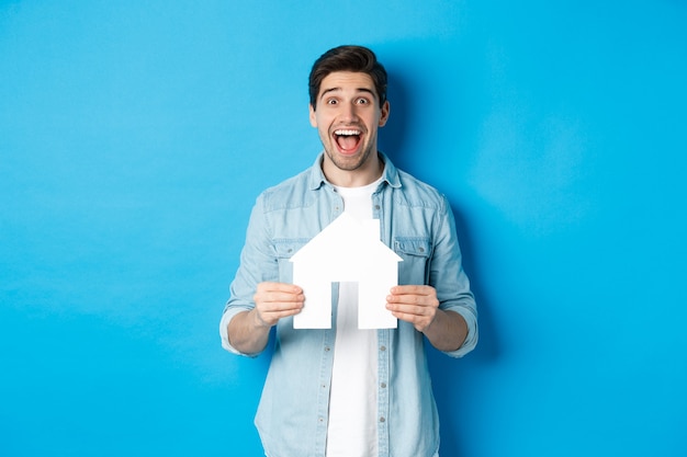 Hombre feliz sosteniendo el modelo de la casa y sonriendo emocionado, comprando una propiedad o alquilando un apartamento, de pie contra la pared azul