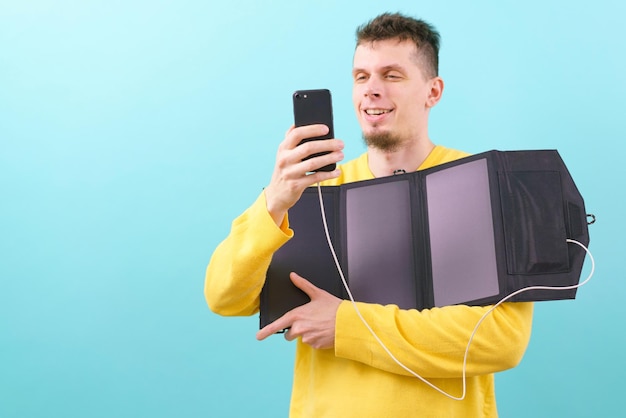 Un hombre feliz sostiene un panel solar portátil y mira su teléfono con un fondo azul Cargador móvil Energía solar verde Eco Electricidad Innovación ambiental Tecnología solar renovable