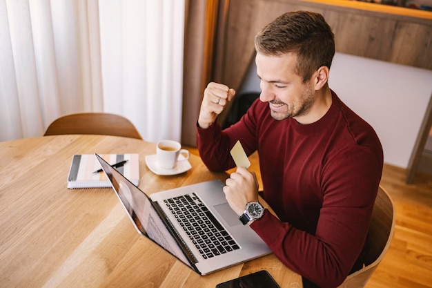 Un hombre feliz tiene una tarjeta de crédito y mira la computadora portátil Un hombre ganó un aumento, por lo que está celebrando