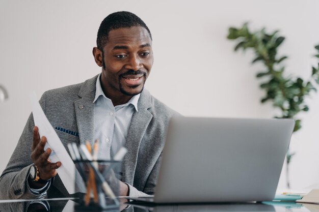 El hombre feliz tiene una videollamada en la computadora portátil y señala documentos de datos Ejecutivo africano remoto