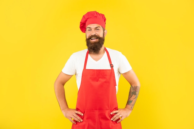 Hombre feliz en toque rojo y delantal con brazos en jarras fondo amarillo, chef