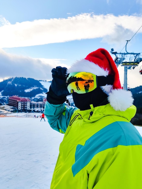 Hombre feliz en traje de esquí con sombrero rojo de navidad de santa en el espacio de copia de la colina de las montañas de invierno