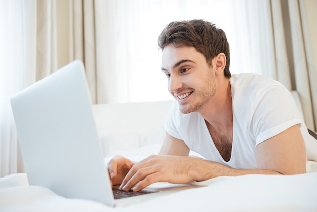 Hombre feliz usando laptop y acostado en la cama. Vista lateral