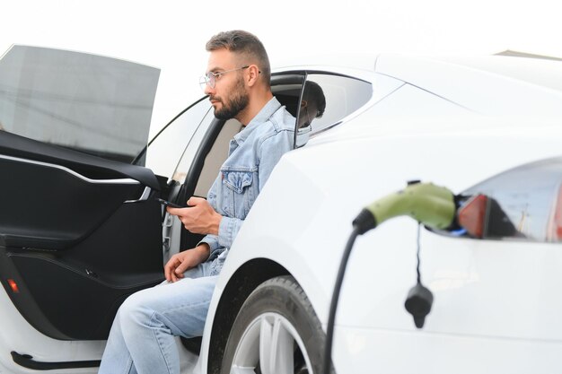 Hombre feliz usando un teléfono inteligente y cargando un coche en una estación de carga de vehículos