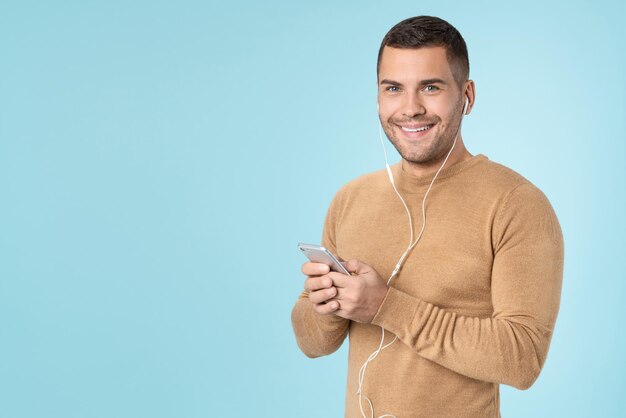 Hombre feliz usando teléfono móvil escuchando música con auriculares posando aislado sobre fondo azul.