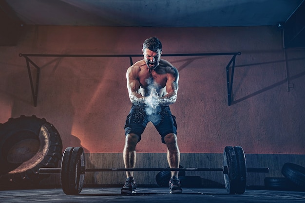 Hombre de fitness muscular que se prepara para hacer peso muerto con una barra sobre su cabeza en el moderno gimnasio. Entrenamiento funcional. Ejercicio de arranque.